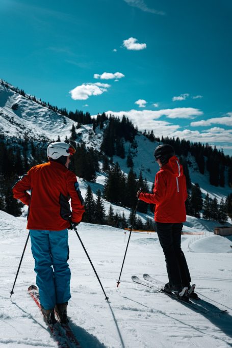 Zwei Skifahrer in roter Kleidung stehen auf einer schneebedeckten Piste mit Bergen im Hintergrund.